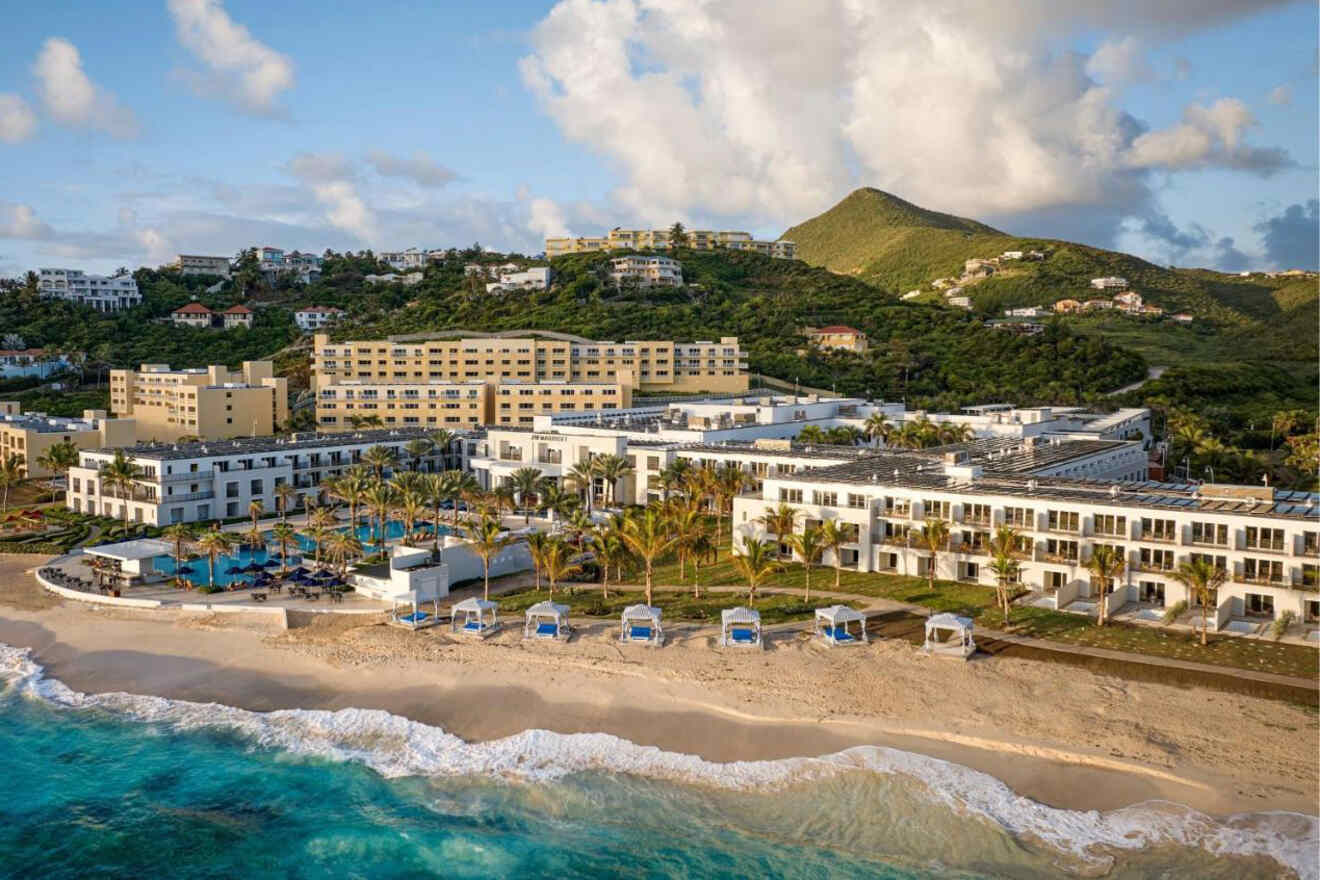 A large beachfront resort with multiple buildings, palm trees, and pools, set against a backdrop of lush green hills and scattered residential homes, with waves gently washing up on the sandy shore.