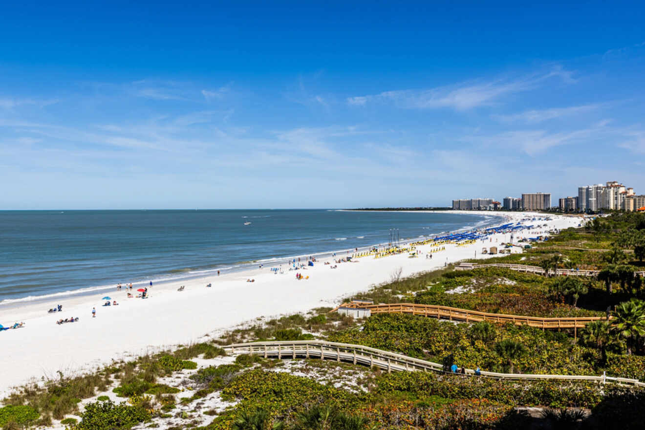 Aerial view of a sandy beach with blue umbrellas, sun loungers, and buildings in the distance under a clear blue sky.