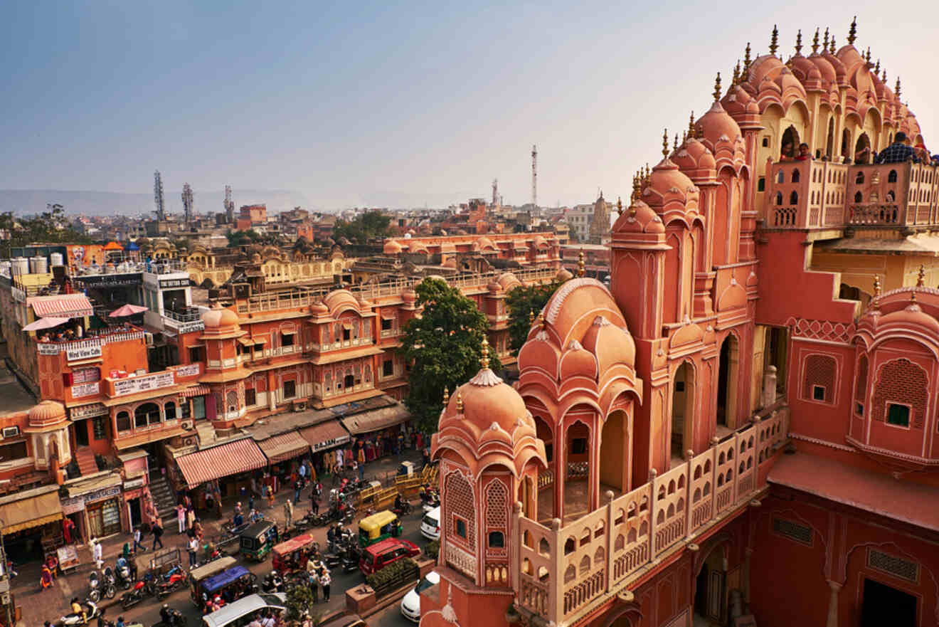 View of Hawa Mahal in Jaipur, with its distinctive pink sandstone facade, overlooking a bustling street filled with vehicles and pedestrians, set against a backdrop of the city's landscape and buildings.