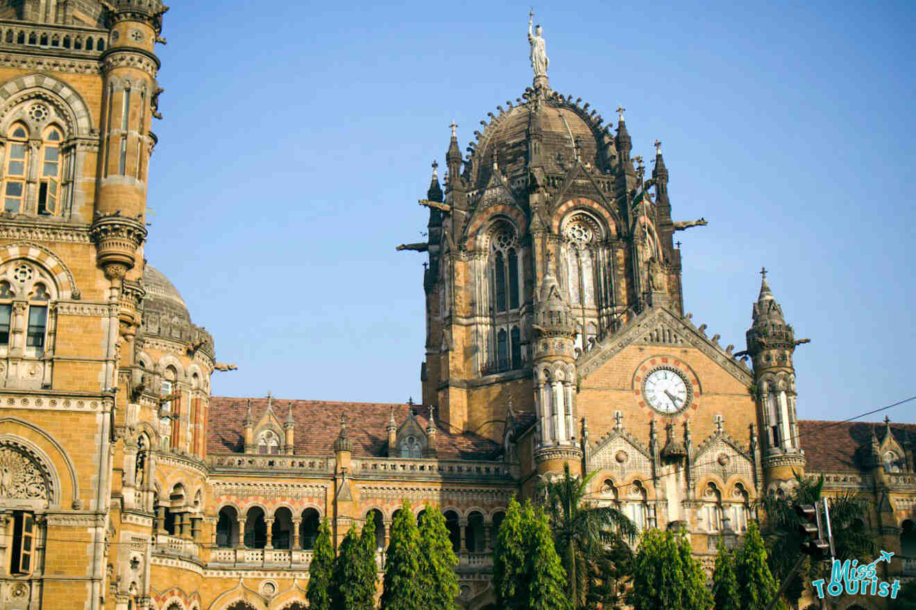 A historic building with ornate Gothic architecture, featuring a central dome, a clock on the facade, and lush green trees in the foreground, set against a clear blue sky.