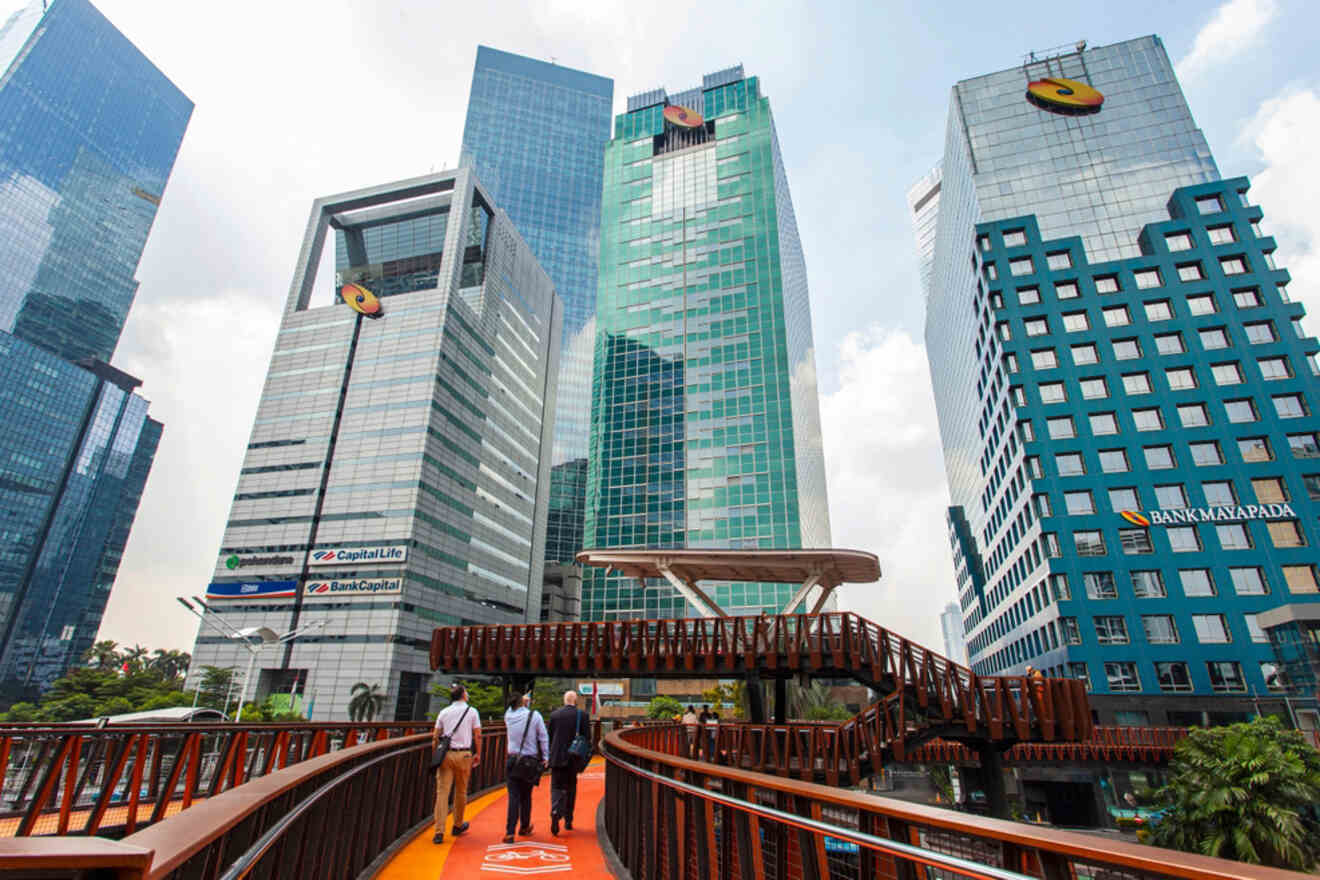 A modern walkway with people crosses between tall, glass skyscrapers on a sunny day. Building logos for CapitaLand and Bank Mandiri are visible.