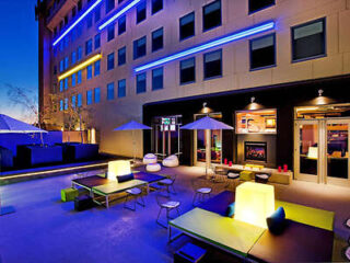 Outdoor patio at night with modern furniture, colorful lighting, and umbrellas. Building facade featuring blue accent lights in the background.