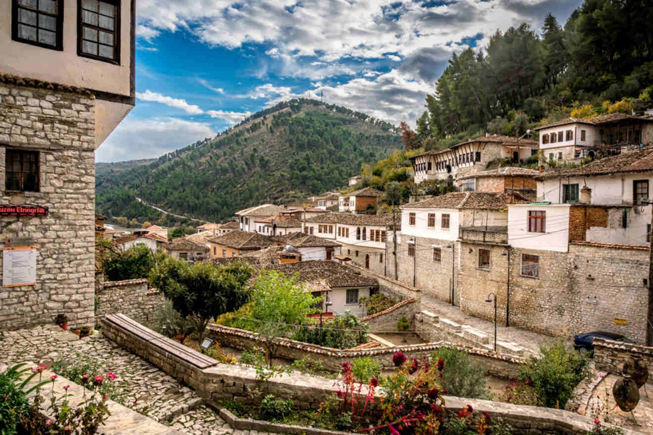 A view of Berat, Albania, with traditional Ottoman-style white houses nestled on the hillside and a green mountain in the background under a partly cloudy sky.