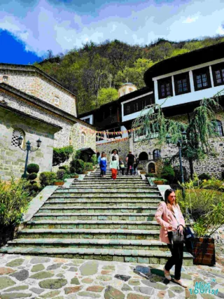 People walking up stone steps surrounded by historical buildings and greenery. The sky is partly cloudy.