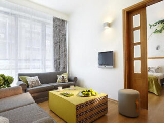 Modern living room with two gray sofas, a green ottoman table, and a wall-mounted TV. A sliding door leads to a bedroom with a botanical artwork.