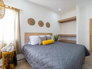 A bedroom with a large bed, gray bedspread, multiple pillows, a yellow bolster pillow, wall decor, open shelving, a tree stump nightstand, rolled towels, and a tall plant in the corner.