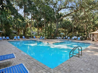 Outdoor swimming pool with surrounding lounge chairs, a small raised platform in the center, and trees in the background.