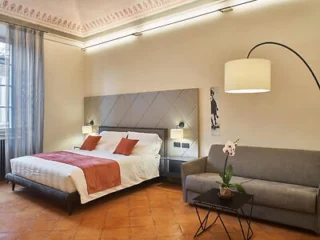 A modern bedroom with a double bed, white linens, orange accents, a gray sofa, a floor lamp, an orchid on a small table, and light streaming in from a window with blue curtains.