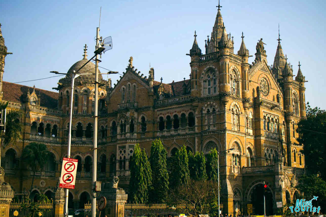 Image shows a large, historic building with Gothic architecture. The building features ornate spires, arched windows, and intricate carvings. There are trees and street signs in the foreground.