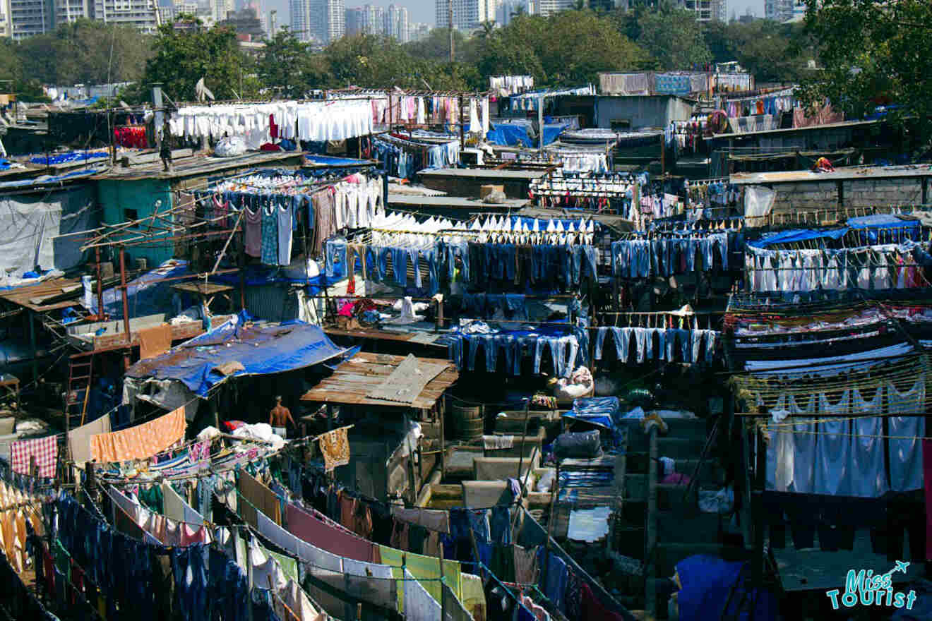 An outdoor laundromat with numerous clotheslines displaying various garments, situated in a densely built urban area with high-rise buildings in the background.