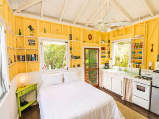 A small, bright yellow room with a double bed, green nightstand, built-in shelves, and a kitchenette featuring a stove, sink, and white cabinets. Large windows allow natural light to stream in.