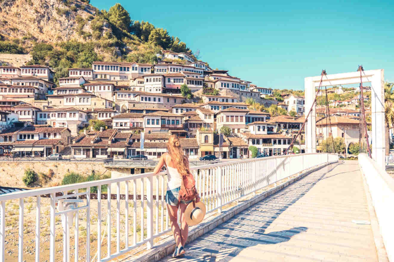 A woman with a backpack and sunhat walks along a bridge in Berat, Albania, overlooking a stunning view of the white, Ottoman-style houses spread across the hillside.