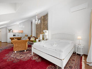 Spacious studio apartment with a white bed, red patterned rug, beige armchairs, and a small dining area under chandeliers, featuring a mix of modern and rustic decor elements.