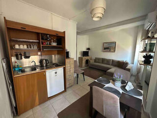 Compact living room and kitchen area with a sofa, TV, dining table set with place settings, and open shelving above a small kitchenette. Neutral tones and a wall-mounted air conditioner are visible.