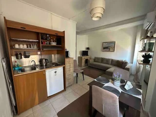 Compact living room and kitchen area with a sofa, TV, dining table set with place settings, and open shelving above a small kitchenette. Neutral tones and a wall-mounted air conditioner are visible.