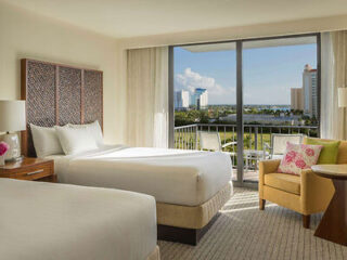 A modern hotel room with two white beds, a plush armchair, and large sliding glass doors