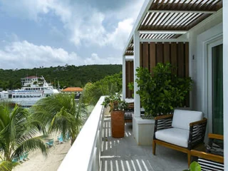 A balcony with a white chair overlooks a tropical scene with palm trees, a beach, and a docked ship. A wooden pergola provides shade overhead.