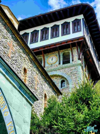 A stone and wood monastery tower with arched windows and a clock face, surrounded by greenery, under a partly cloudy sky.