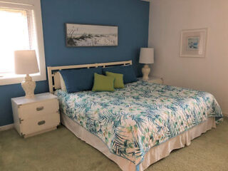 A bedroom with a queen-size bed, light blue and green bedspread, two matching bedside tables with lamps, blue accent wall, and framed artwork above the bed.