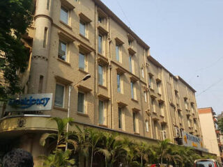 A multistory beige brick building with several windows, surrounded by palm trees. Signs on the building read "Residency" and "Abhinav.