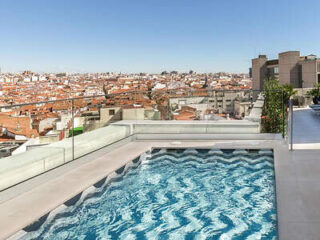 Rooftop swimming pool with clear water, surrounded by glass barriers, overlooking a cityscape with numerous buildings under a blue sky.