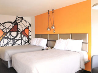 A hotel room with two double beds, white bedding, an orange accent wall, and modern black and orange wall art.