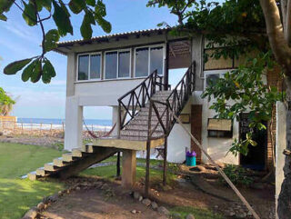 A house on stilts with an external staircase leading to a second-floor balcony. The building is near the beach, surrounded by greenery and trees.