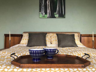 A neatly made bed with yellow and white bedding, two dark brown pillows, and a wooden tray holding two blue mugs. The wall behind the bed has a painting of tall trees.