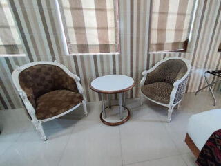 Two upholstered chairs with patterned fabric flank a small round table in a room with striped window blinds and light-colored tile flooring.