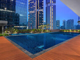 A swimming pool with a wooden deck is surrounded by tall modern buildings in a cityscape at dusk.