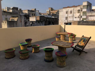 Rooftop with traditional woven stools, two woven tables, and a decorative chair under clear sky. Background of urban rooftops with old buildings.