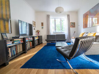 A modern living room with black furniture, a blue rug, a large TV, bookshelves, and a window letting in natural light. A guitar leans against the wall near the window.