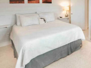 A neatly made bed with white linens, two pillows, and a bedside lamp in a minimalistic bedroom.