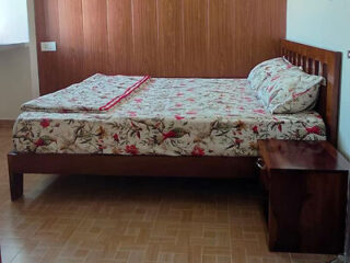 A neatly made bed with floral patterned sheets and pillows, situated in a room with wooden flooring and a wooden paneled wall. A small wooden nightstand is placed next to the bed.