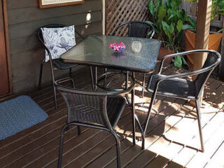 A glass-top table with four black chairs is set on a wooden deck. A small vase with pink flowers is centered on the table.