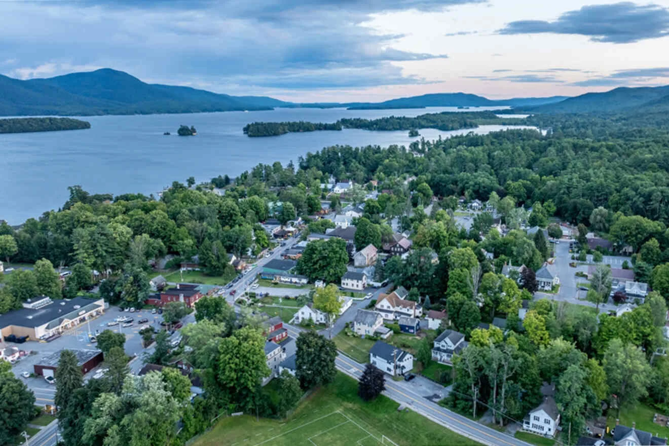 Aerial view of a small town with surrounding greenery and a large body of water in the background, under a partly cloudy sky.