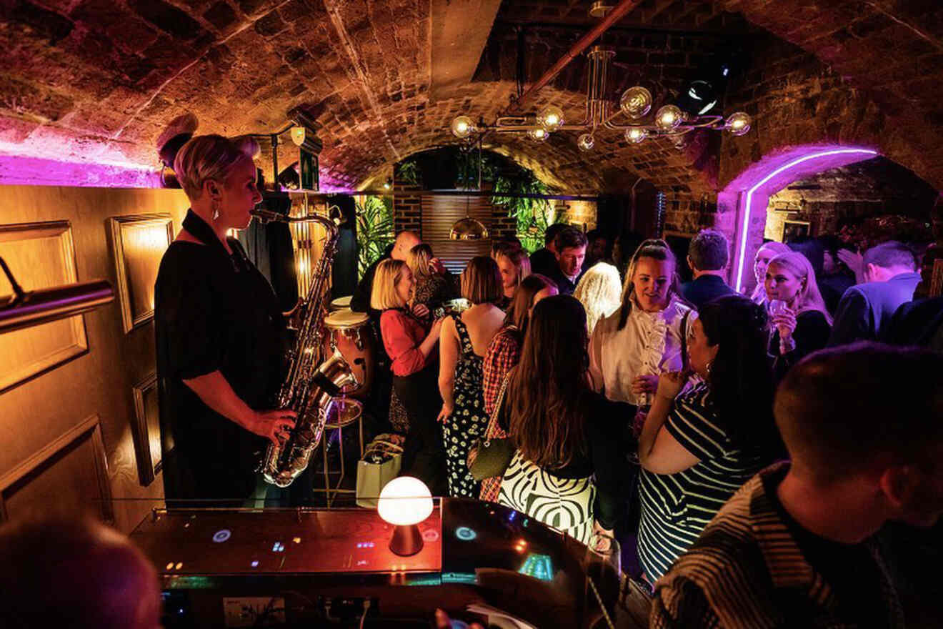 A saxophonist plays live music to a lively crowd under a low ceiling with purple lighting, creating an intimate party atmosphere.