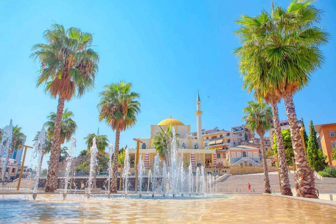 A mosque with a yellow dome and a minaret is seen in the background, framed by several tall palm trees and a water fountain in the foreground under a clear blue sky.