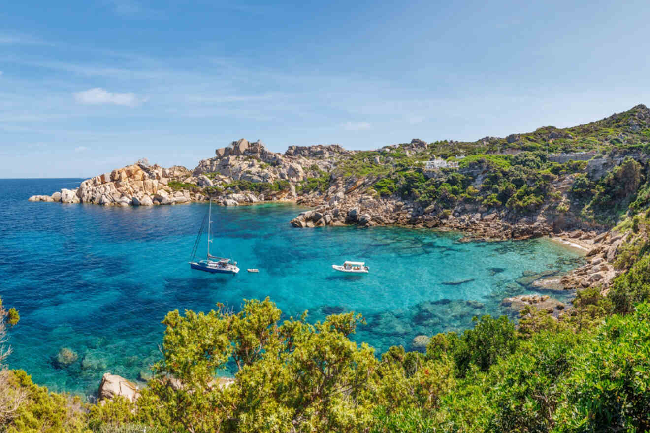 A picturesque cove with clear blue waters, sailboats anchored near rocky shores, and dense greenery surrounding the beach area.