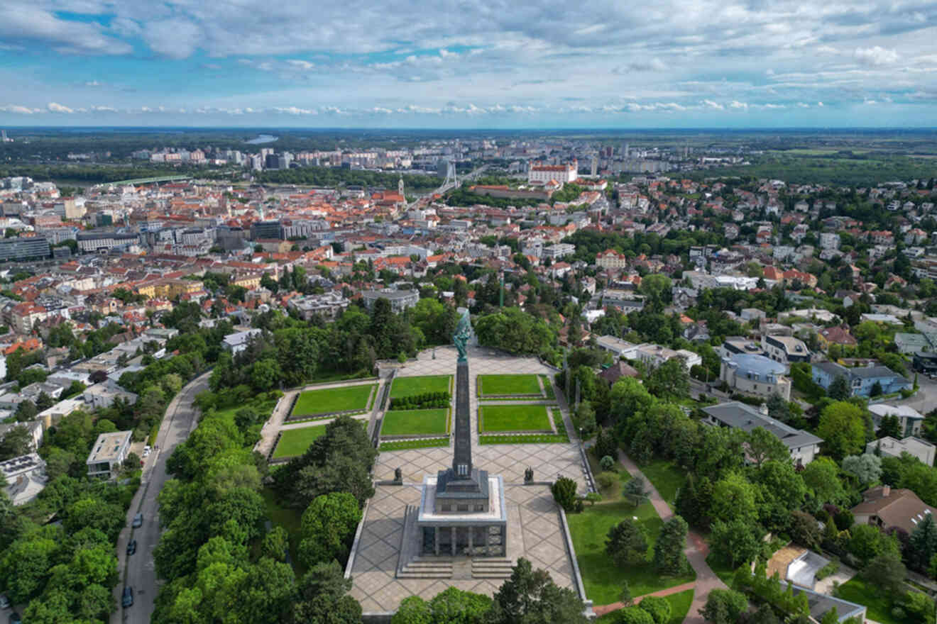 Aerial view of a cityscape with a prominent monument centered in a large park surrounded by greenery, residential buildings, and an expansive urban area extending into the distance.