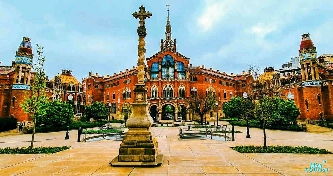 A historic brick building with arched windows and ornate stone details, surrounded by trees and a courtyard featuring a statue and fountain, under a cloudy sky.