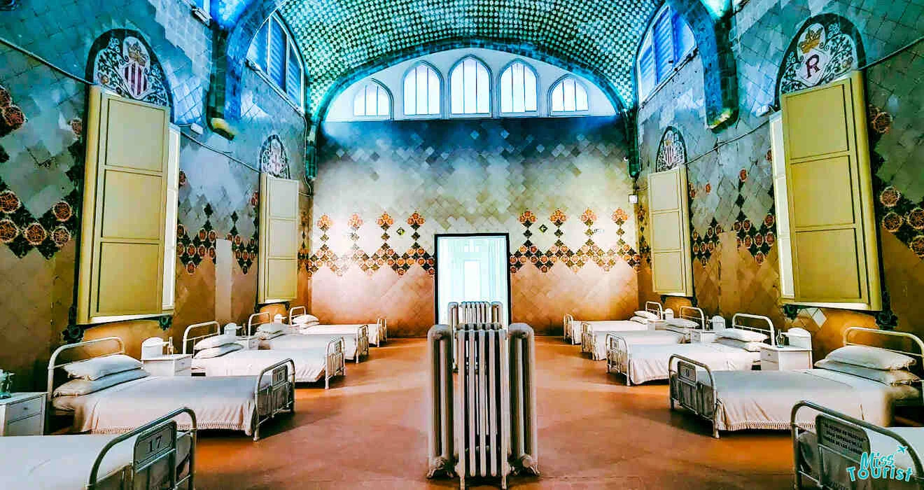 A spacious room with several metal-framed hospital beds arranged in rows, a central screen divider, and walls featuring colorful tiled patterns under a high, arched ceiling with decorative windows.