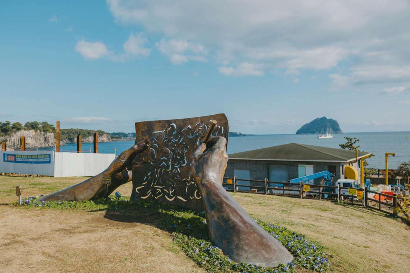 A large sculpture of two hands holding an open book with intricate designs is situated on a grassy area near the sea with a small building and an island in the background.