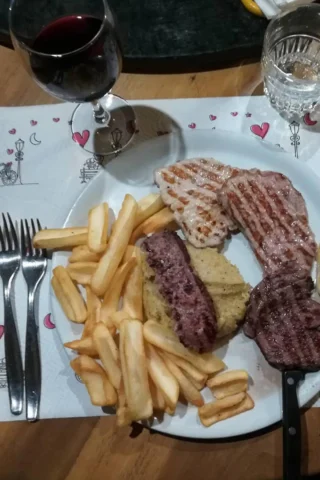 A plate with grilled meats, fries, and polenta. A glass of red wine and a glass of water are placed above the plate. Two forks and a knife are beside the plate on a decorated placemat.