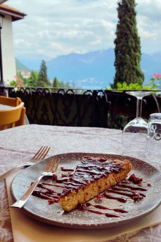 A piece of cake drizzled with chocolate sauce on a plate is set on a table with cutlery and a glass, overlooking a scenic mountain and nature view.