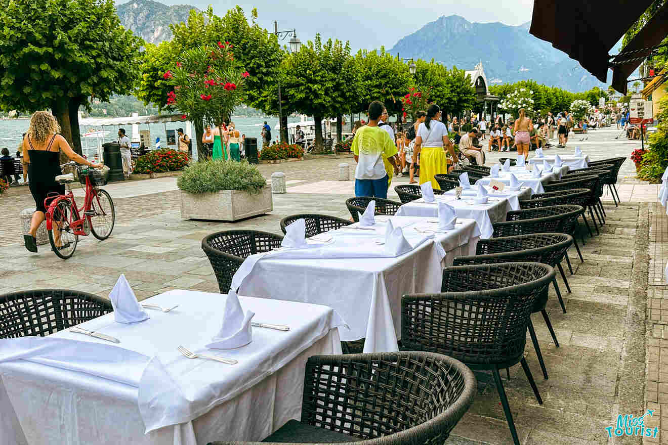 Outdoor dining area with tables set with white tablecloths and napkins. People walk and cycle along a pedestrian path lined with trees, mountains visible in the background.