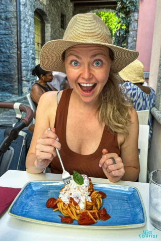 author of the post wearing a wide-brimmed hat is sitting at an outdoor table, looking surprised while holding a fork near a plate of pasta topped with cheese and basil.