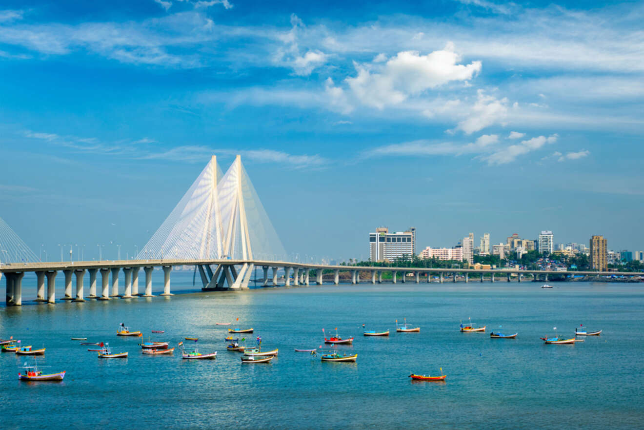 Image of a cable-stayed bridge over a body of water, with numerous small boats and a skyline of buildings in the background under a clear blue sky.