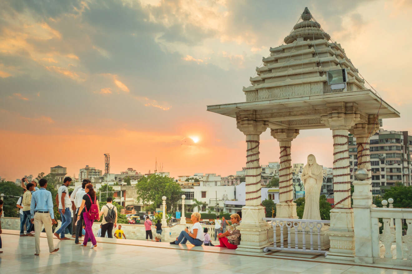 People gather at a temple terrace during sunset, with a statue and a pillared structure in the foreground and a cityscape in the background.