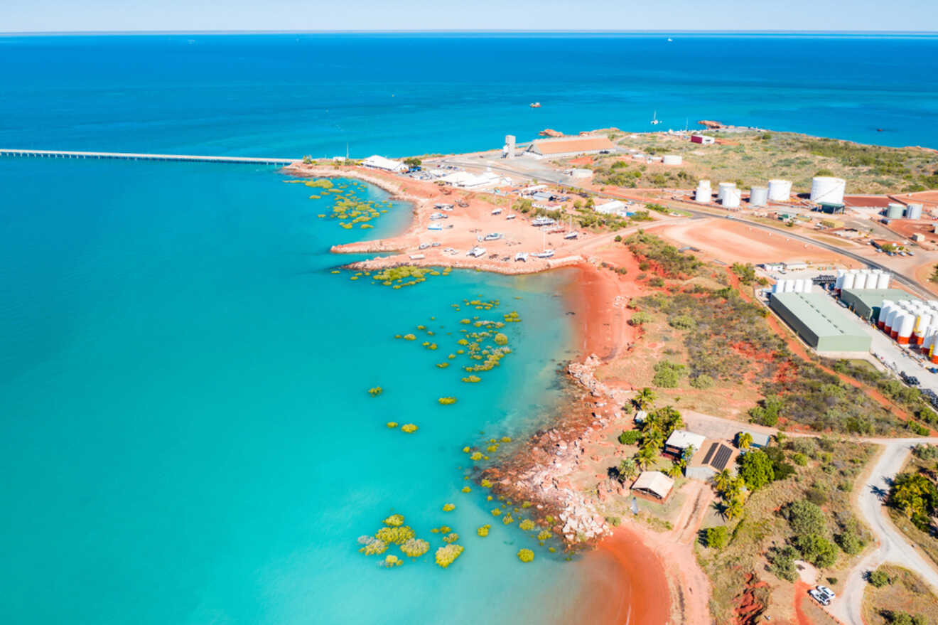 Aerial view of a coastal landscape featuring turquoise waters, coral formations, industrial buildings, and storage tanks near a shoreline with red soil and green vegetation.